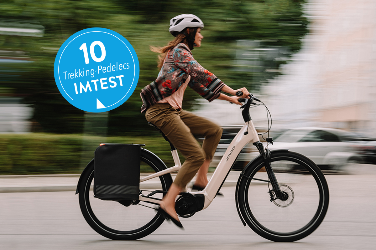 Frau fährt auf weißem Trekking-E-Bike in der Stadt; links ein blauer Sticker mit 