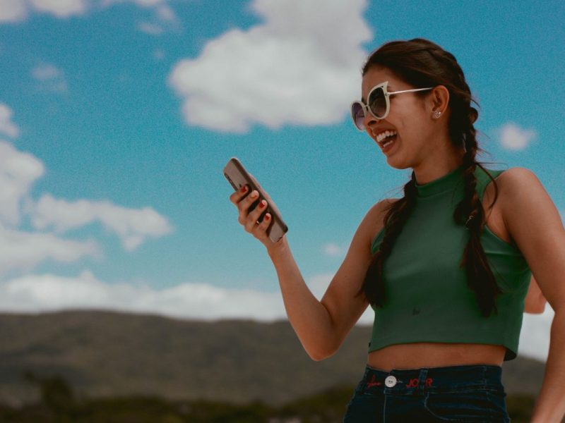Eine Frau im grünen Tank Top mit weißer Sonnenbrille schaut lachend auf ihr Smartphone. Im Hintergrund ein heiterer Himmel und Berge.