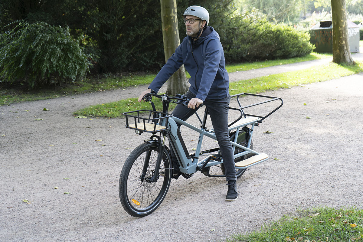 Mann sitzt auf Decathlon Cargobike R500E Langtail und hat den linken Fuß auf dem Boden vor Parklandschaft abgestellt.