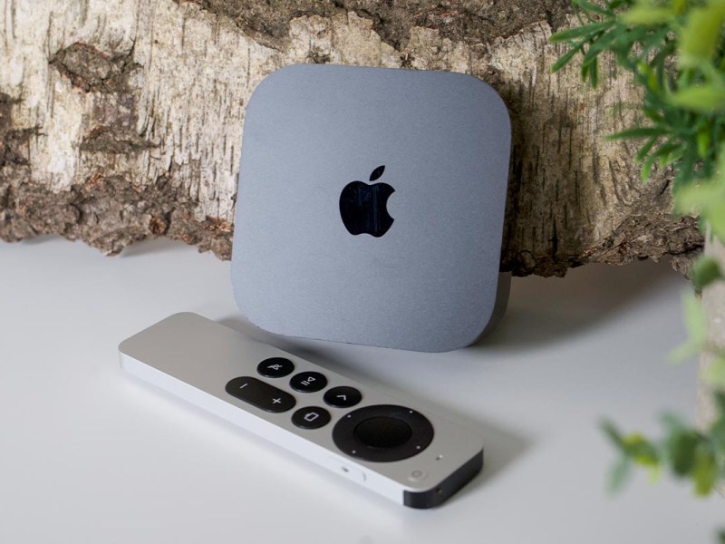 Apple TV 4K mit Fernbedienung vor einem Holzstück auf weißem Untergrund.