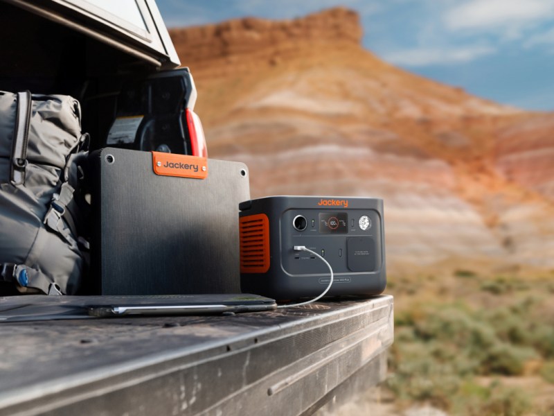 Kleine orange schwarze Jackery Powerstation mit angeschlossenem Smartphone auf offenem Autoheck vor Wüstenkulisse