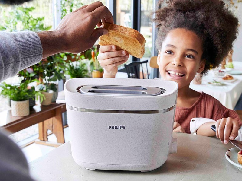 Kleines Mädchen lächelt, während es eine Scheibe Toast über die Schlitze des Toasters hält. Die Hand eines Mannes greift von vorn ins Bild und fasst die Brotscheibe an der linken Ecke an