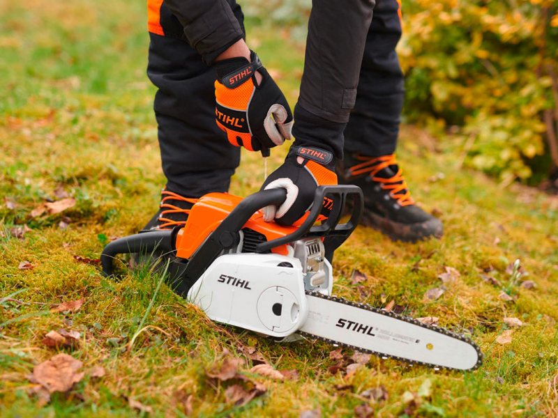 Benzin-Kettensäge von Stihl, auf einer Wiese, dahinter ein Menschen in schwarz-oranger Sicherheitskleidung, der versucht, die Kettensäge zu starten.