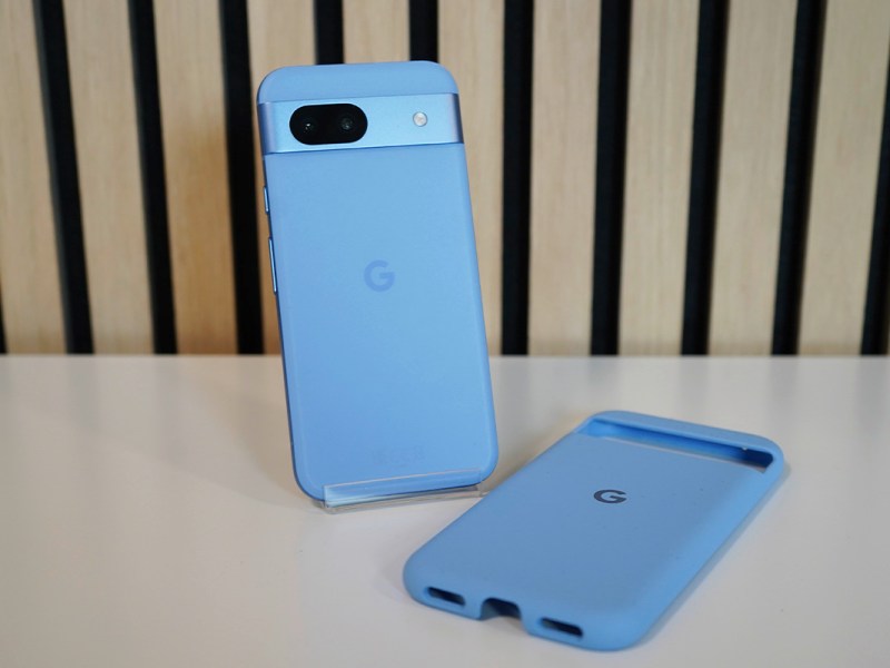 Ein hellblaues Smartphone auf einem weißen Tisch vor einer Wand mit heller Holzverkleidung.