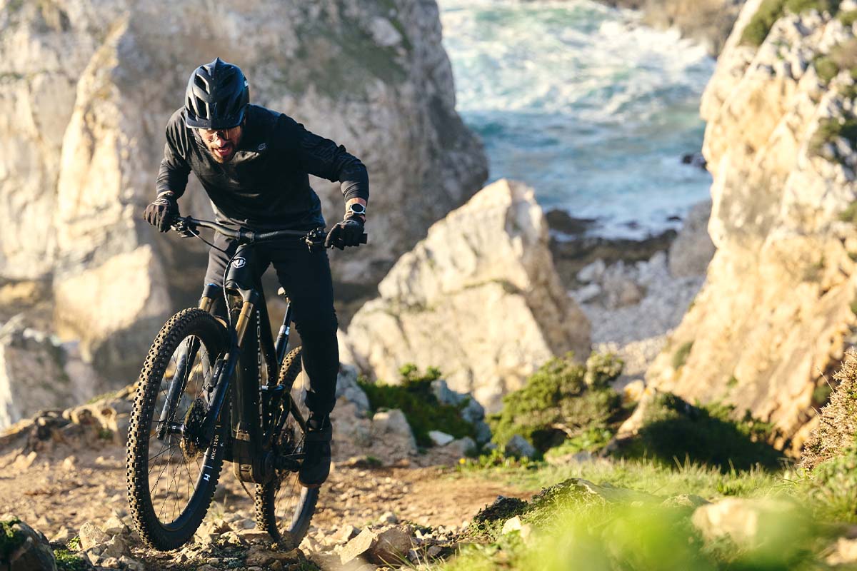 Mann fährt mit einem E-MTB einen steilen Trail hoch, Meer und Felsen im Hintergrund